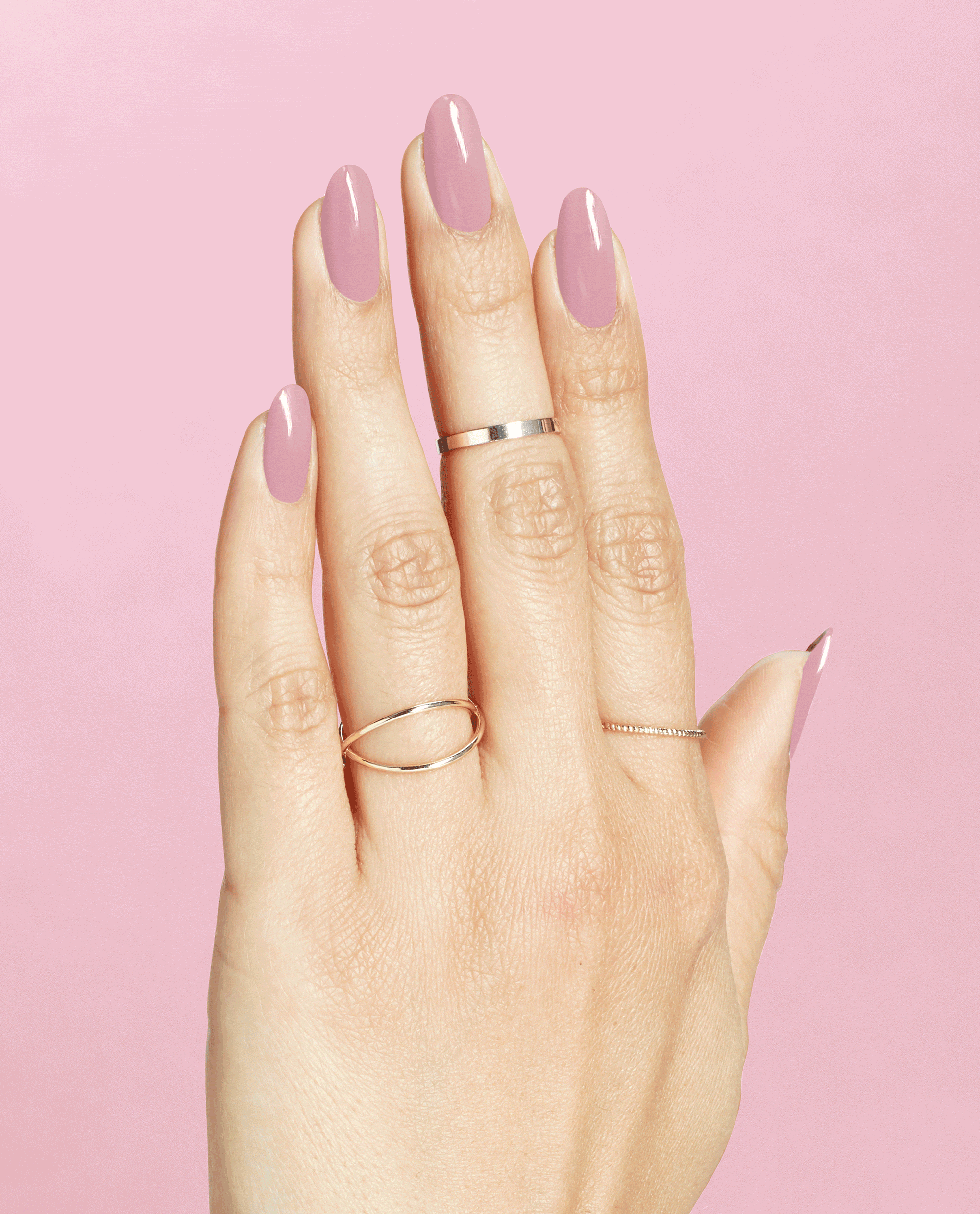 Hand with long oval nails painted in (P)Ink on Canvas, a glossy mauve-pink OPI nail polish, resting against a solid pink background and styled with delicate gold and silver rings to showcase the nail lacquer color and shine.