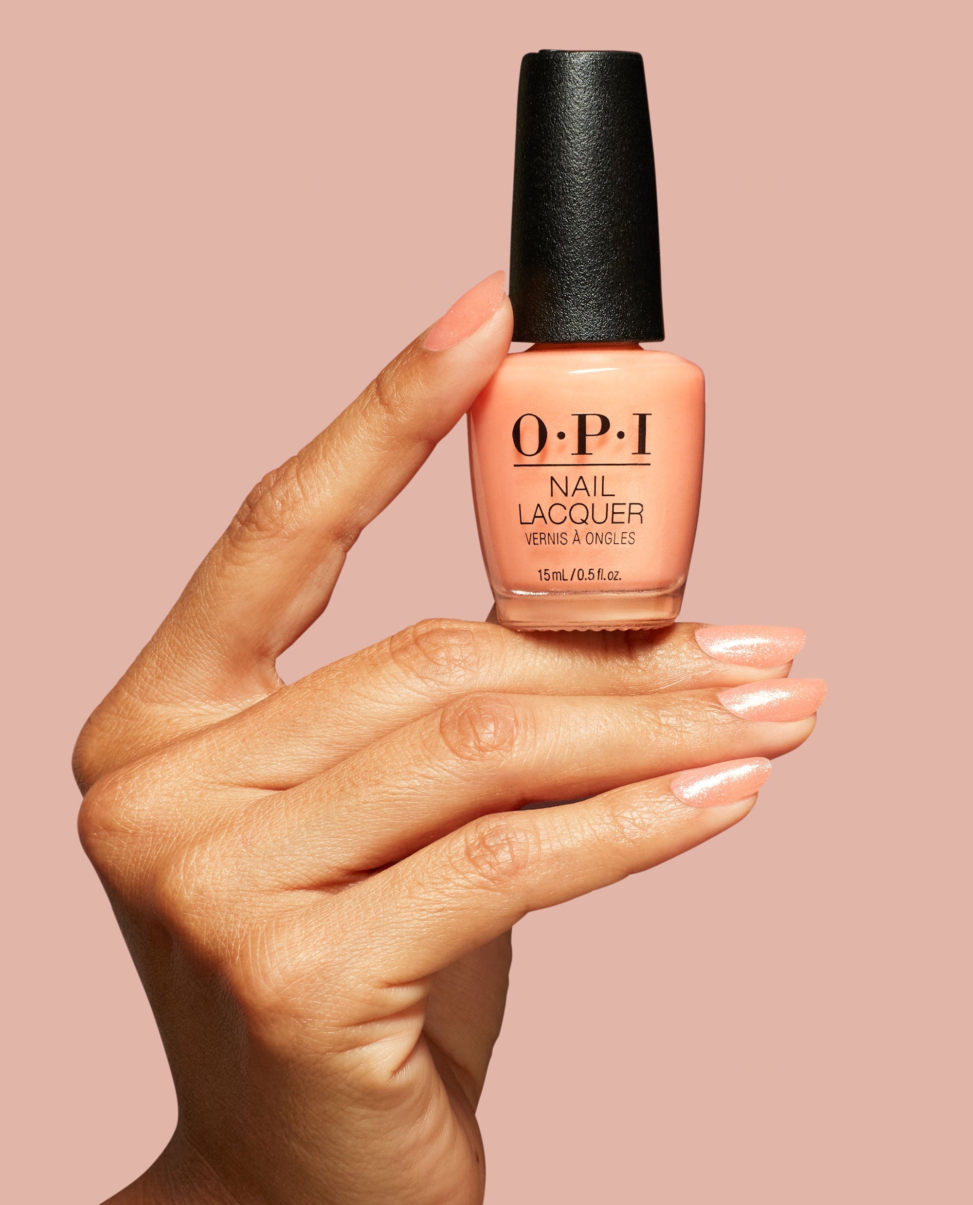 Hand with glossy peach shimmer nails holds OPI Queens Rule Nail Lacquer bottle, a trending soft coral shade with black cap, against a blush pink background.