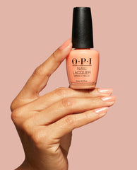 Hand with glossy peach shimmer nails holds OPI Queens Rule Nail Lacquer bottle, a trending soft coral shade with black cap, against a blush pink background.