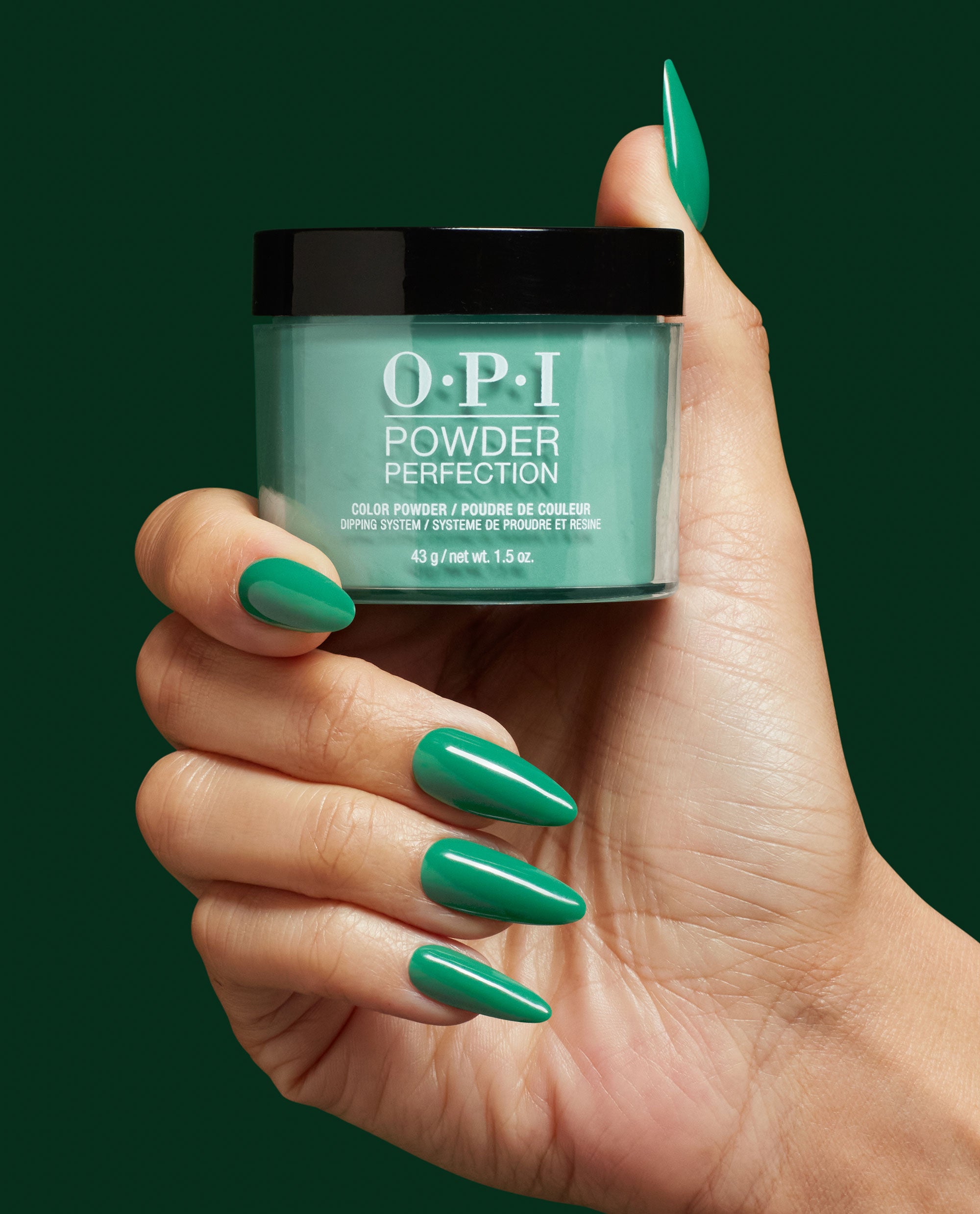 Rated Pea-G OPI Powder Perfection dipping powder jar held by a hand with long almond nails painted glossy emerald, showcasing green nail polish ideal for spring nail colors, with OPI branding on the mint jar against a dark green backdrop.