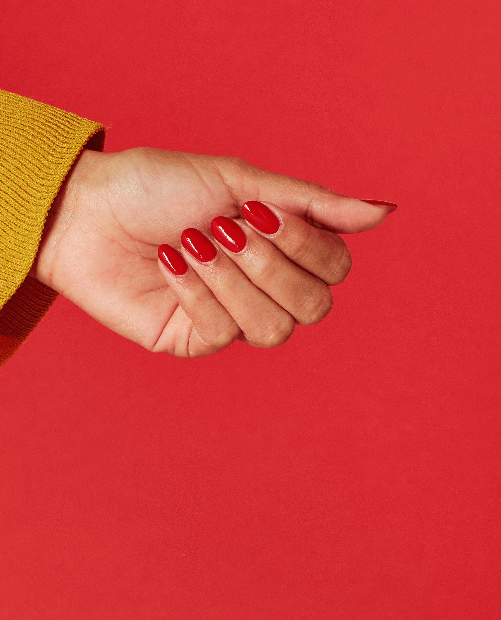 Hand with oval-shaped nails painted in glossy bright red Red Heads Ahead OPI nail polish, shown against a solid red background with a mustard-yellow sweater cuff highlighting the bold nail lacquer color.