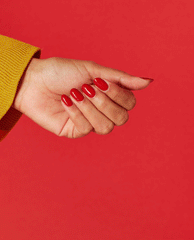 Hand with oval-shaped nails painted in glossy bright red Red Heads Ahead OPI nail polish, shown against a solid red background with a mustard-yellow sweater cuff highlighting the bold nail lacquer color.