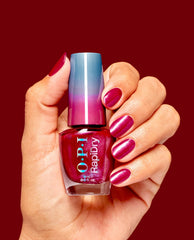 Hand holding OPI RapiDry Ring the Alarm nail polish, a shimmery magenta-red fall nail color, with matching glossy manicured nails against a deep red background.