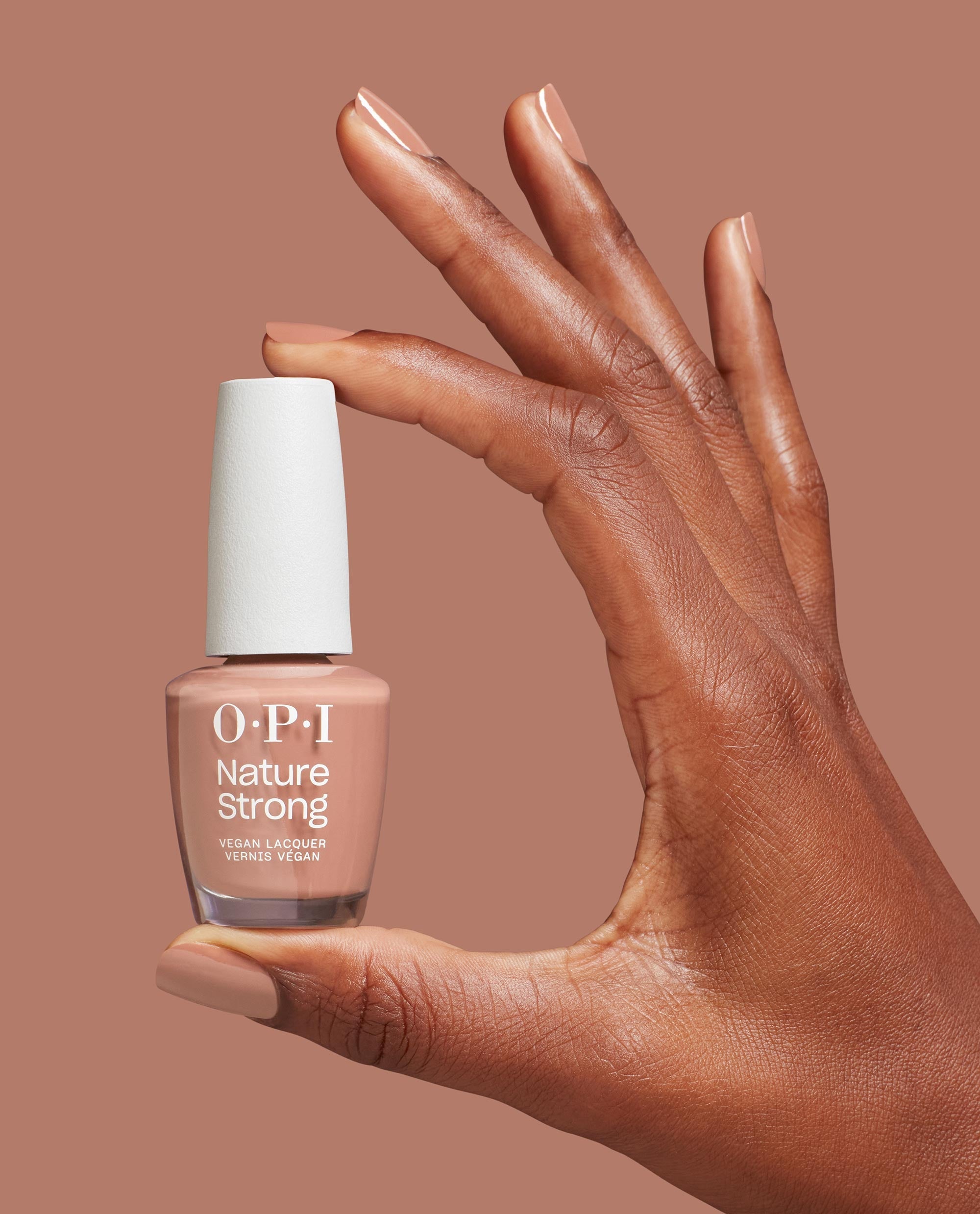 Hand with glossy nude-pink nails holds OPI Nature Strong Vegan Nail Lacquer in Rooting For Hue, a trending vegan nail polish shade, against a warm mauve background.