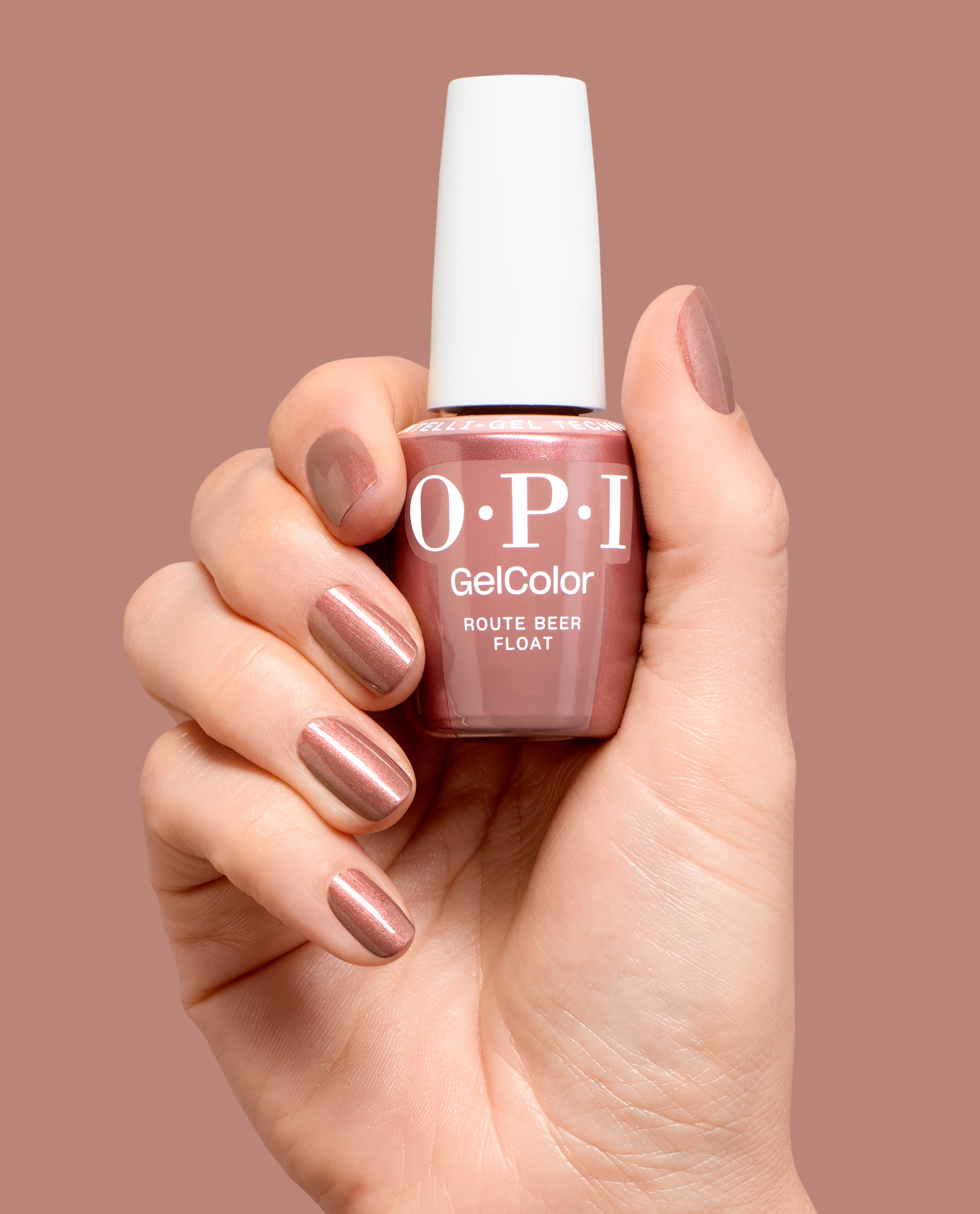 Hand holds OPI GelColor Route Beer Float gel nail polish in a shimmery rosy brown shade, with matching manicured nails against a mauve backdrop for fall nail colors.