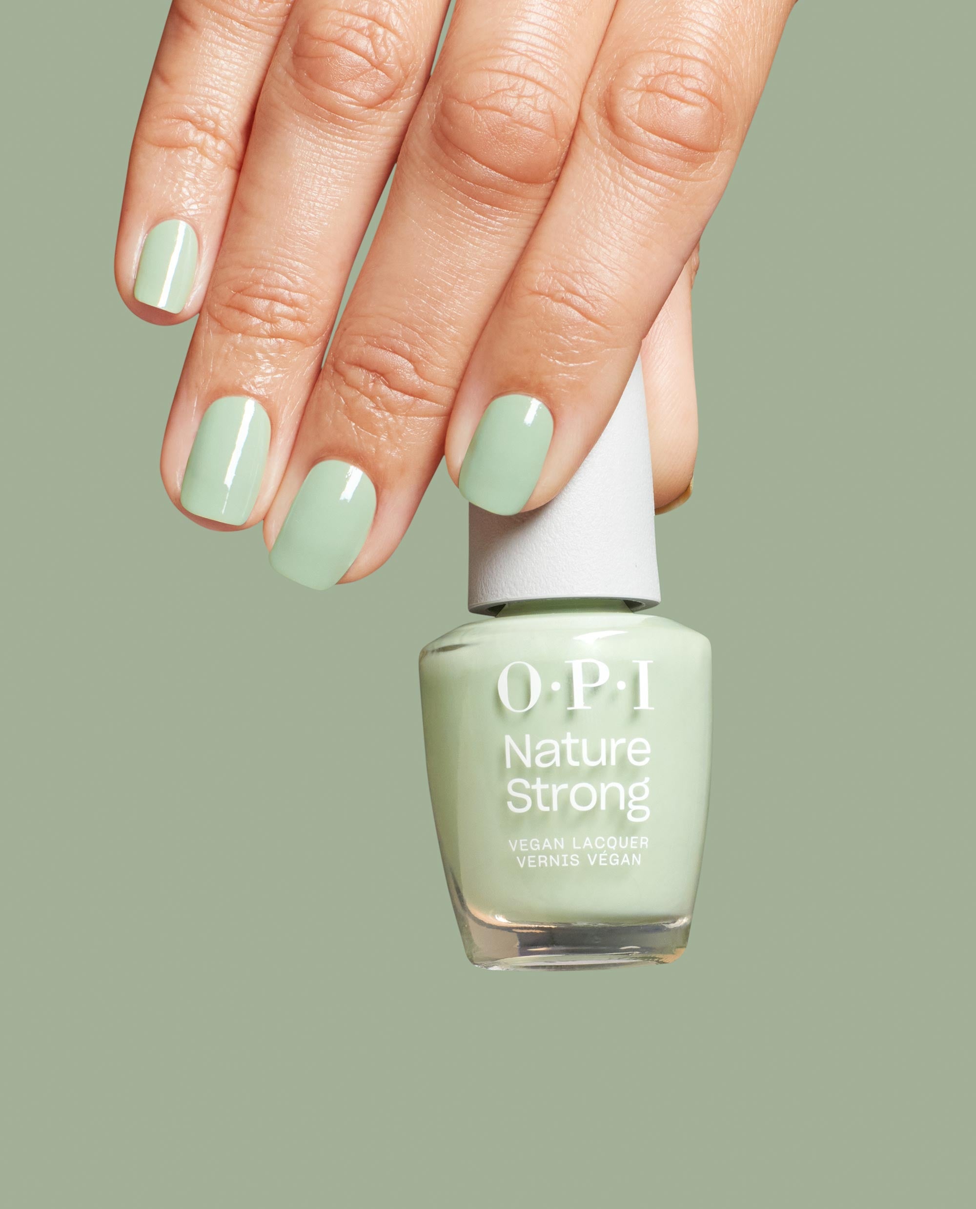 Hand with glossy sage green nails holds OPI Nature Strong Vegan Nail Lacquer in Sage It for Later, matching the pale green polish bottle against a soft sage background.