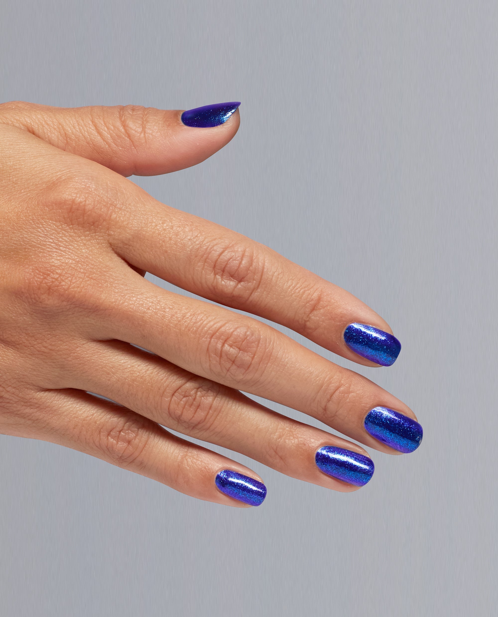 Hand models shimmering deep blue Scorpio Seduction OPI nail polish nail lacquer on neatly manicured nails, showing glossy, glittery coverage against a soft gray background for bold, celestial-inspired manicure look.