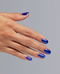 Hand models shimmering deep blue Scorpio Seduction OPI nail polish nail lacquer on neatly manicured nails, showing glossy, glittery coverage against a soft gray background for bold, celestial-inspired manicure look.