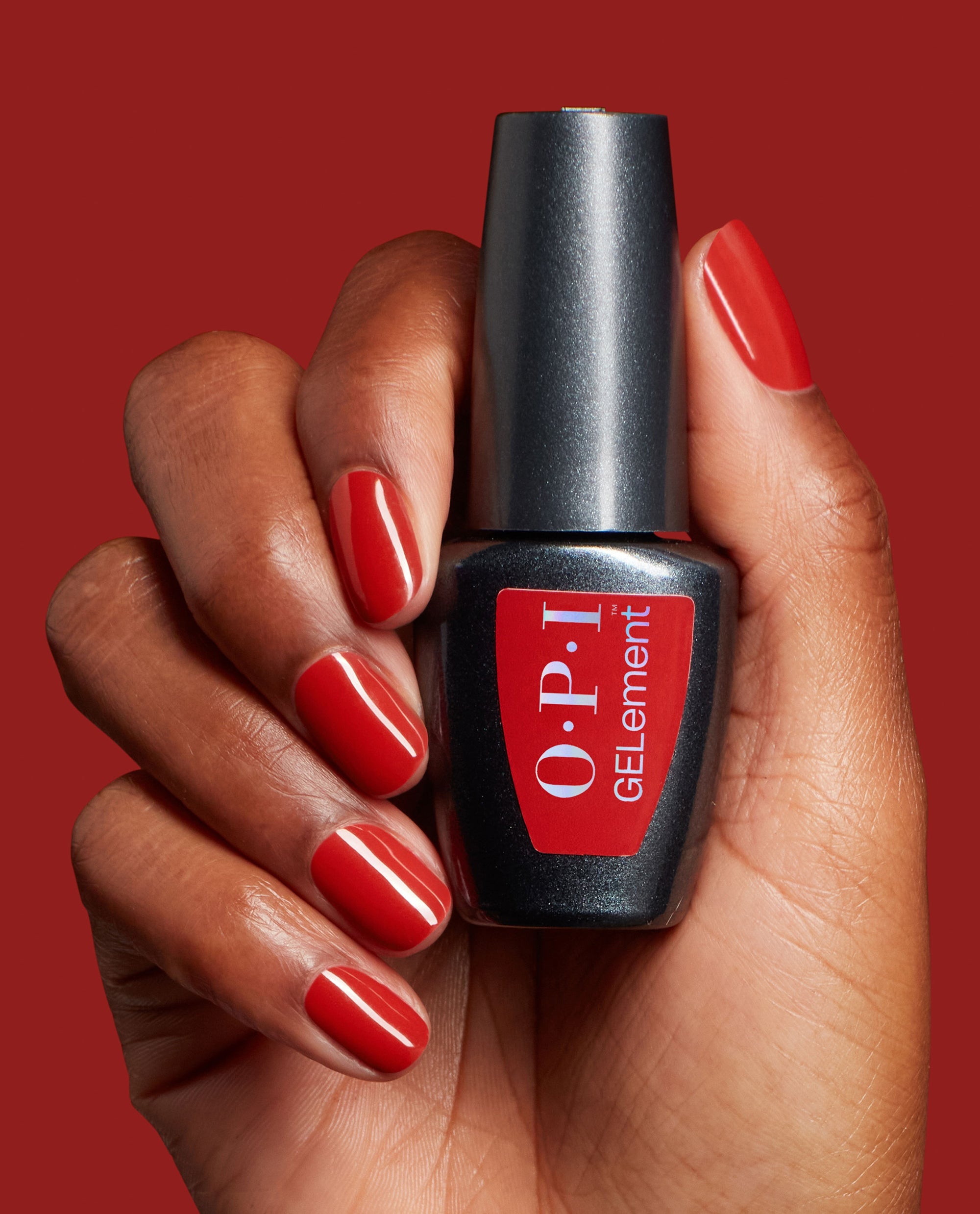 Hand with glossy red gel manicure holds OPI GelColor Single & Red-y at-home gel nail polish against a red background, a bold choice for fall nail colors.