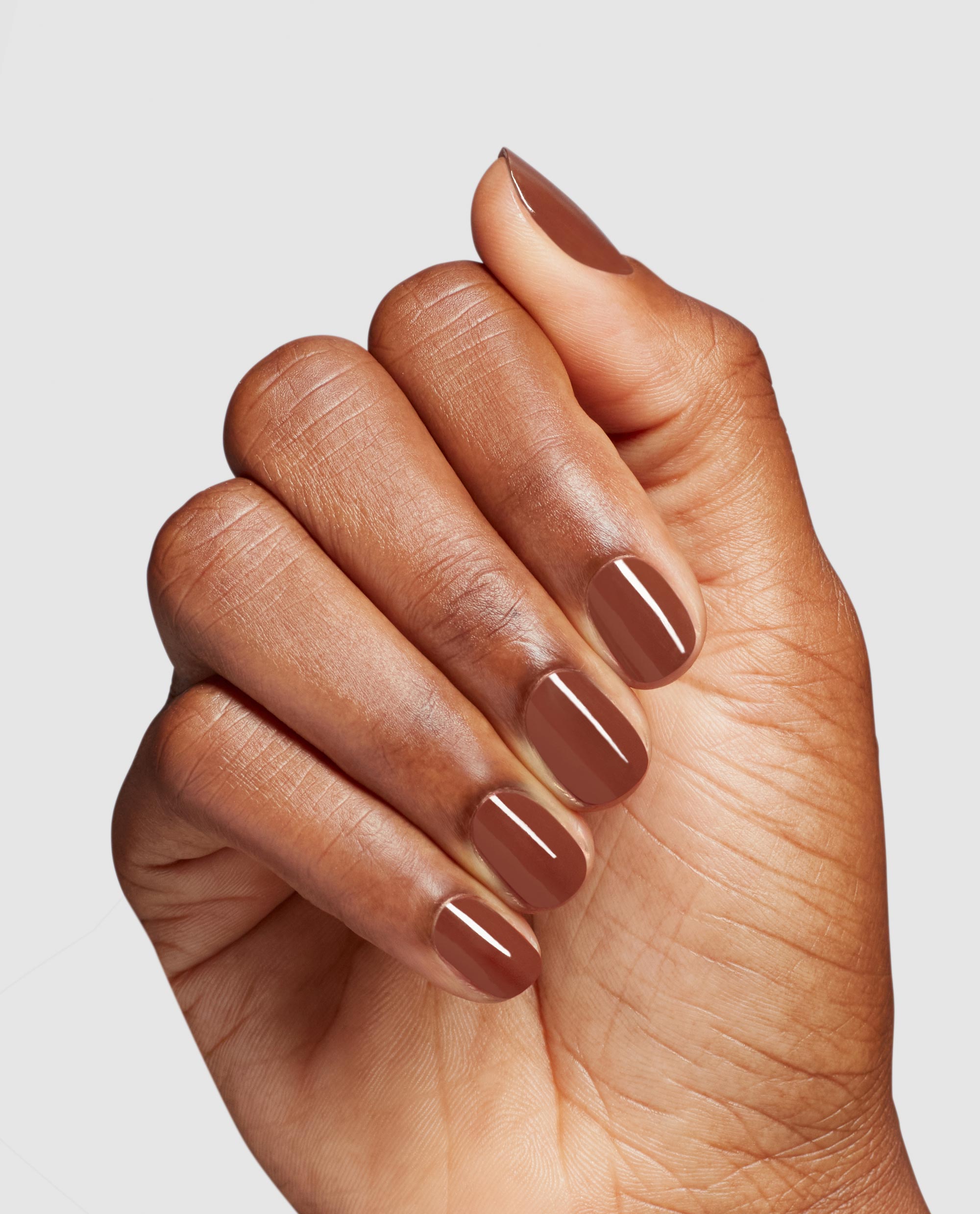 Hand displays glossy OPI Slip Dressed Up nail lacquer, a warm brown fall nail color on short rounded nails against a clean light gray studio background.