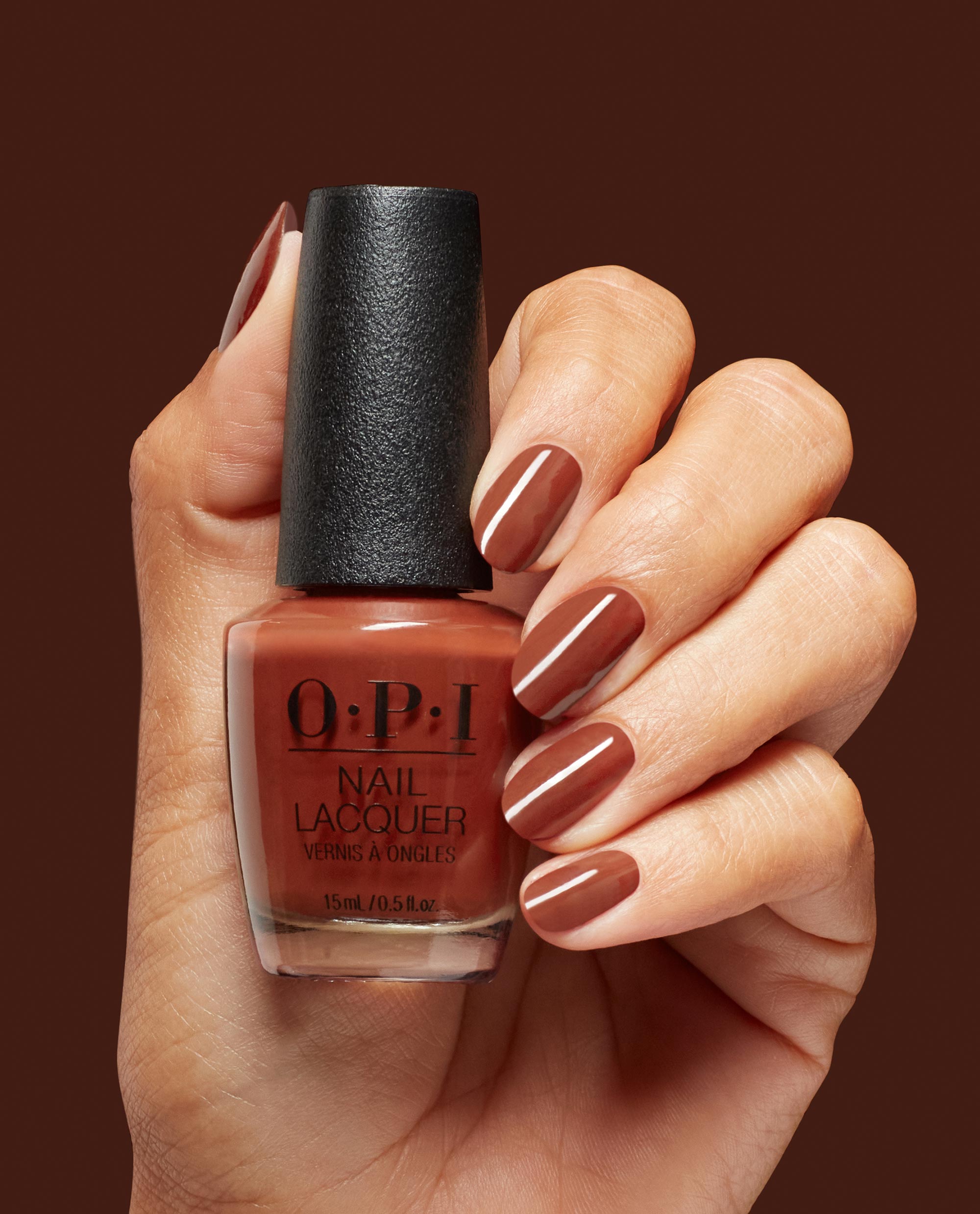 Hand holding OPI Slip Dressed Up Nail Lacquer, showing glossy warm brown manicured nails that match the bottle shade against a rich brown background, ideal for fall nail colors.