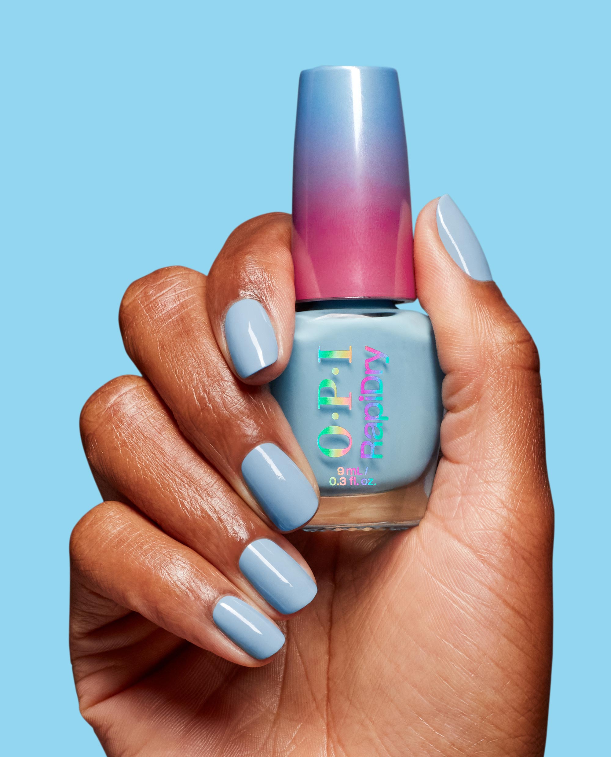 Hand with glossy light blue manicure holds OPI Snap Your Fingers RapiDry nail polish bottle with blue-to-pink ombre cap against a bright blue background, showcasing the nail polish gift set color and sleek product packaging.