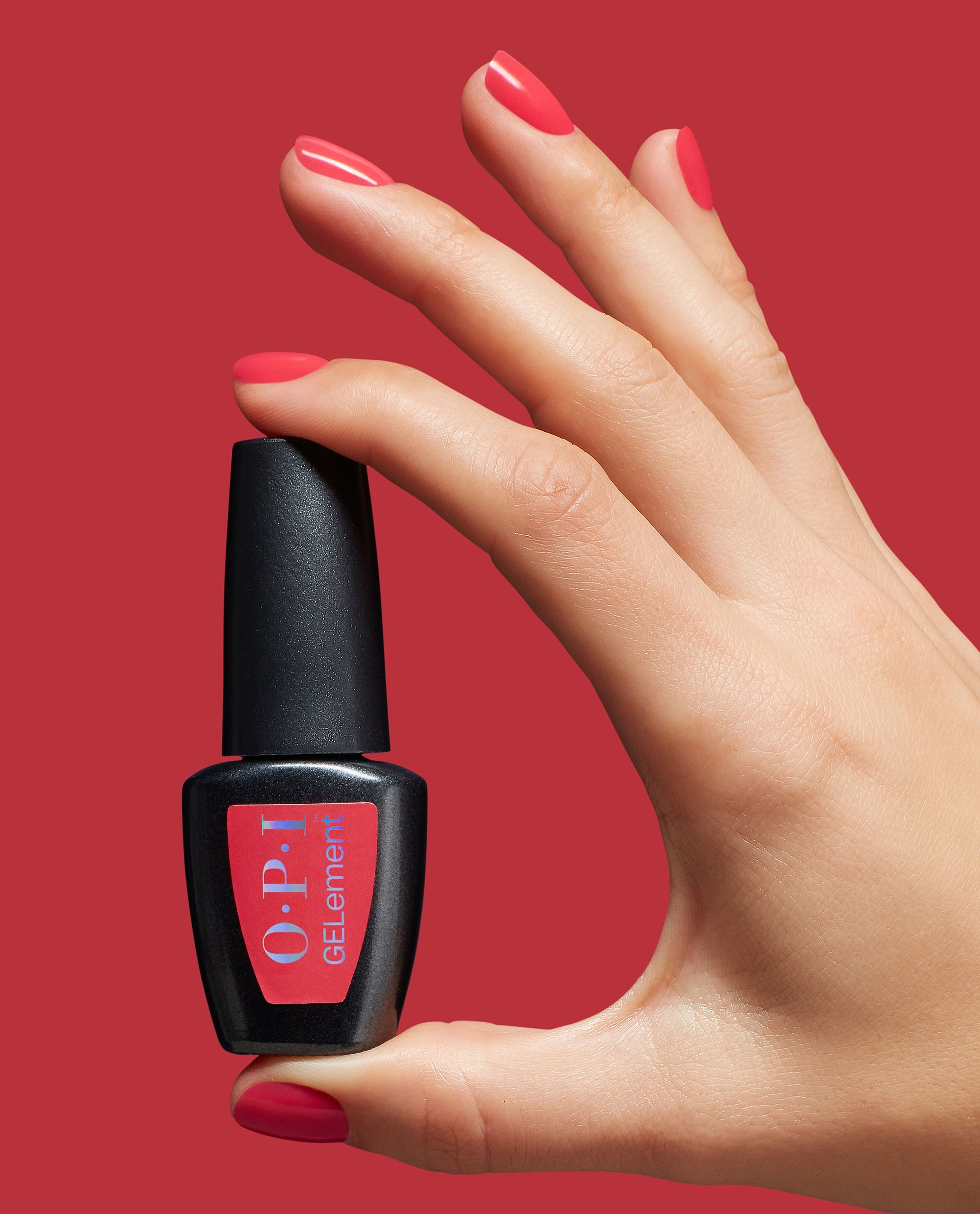 Hand with glossy coral-red nails holds OPI GelColor Special O-Cajun at-home gel nail polish bottle against a matching red background, showcasing a trending manicure shade.