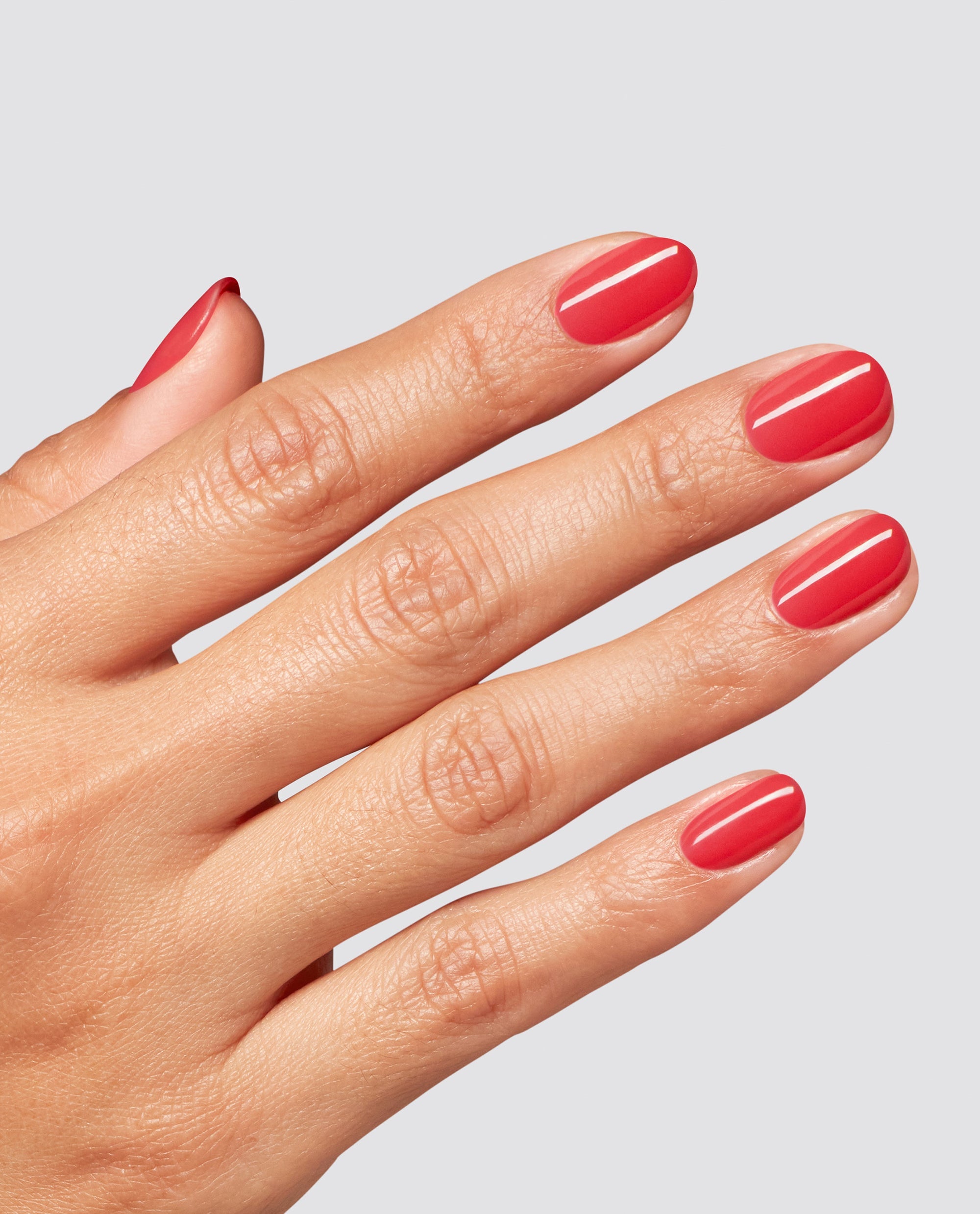 Hand shows glossy coral-red Special O-Cajun at-home gel nail polish on short rounded nails against a light gray background, a trending OPI-inspired manicure shade.