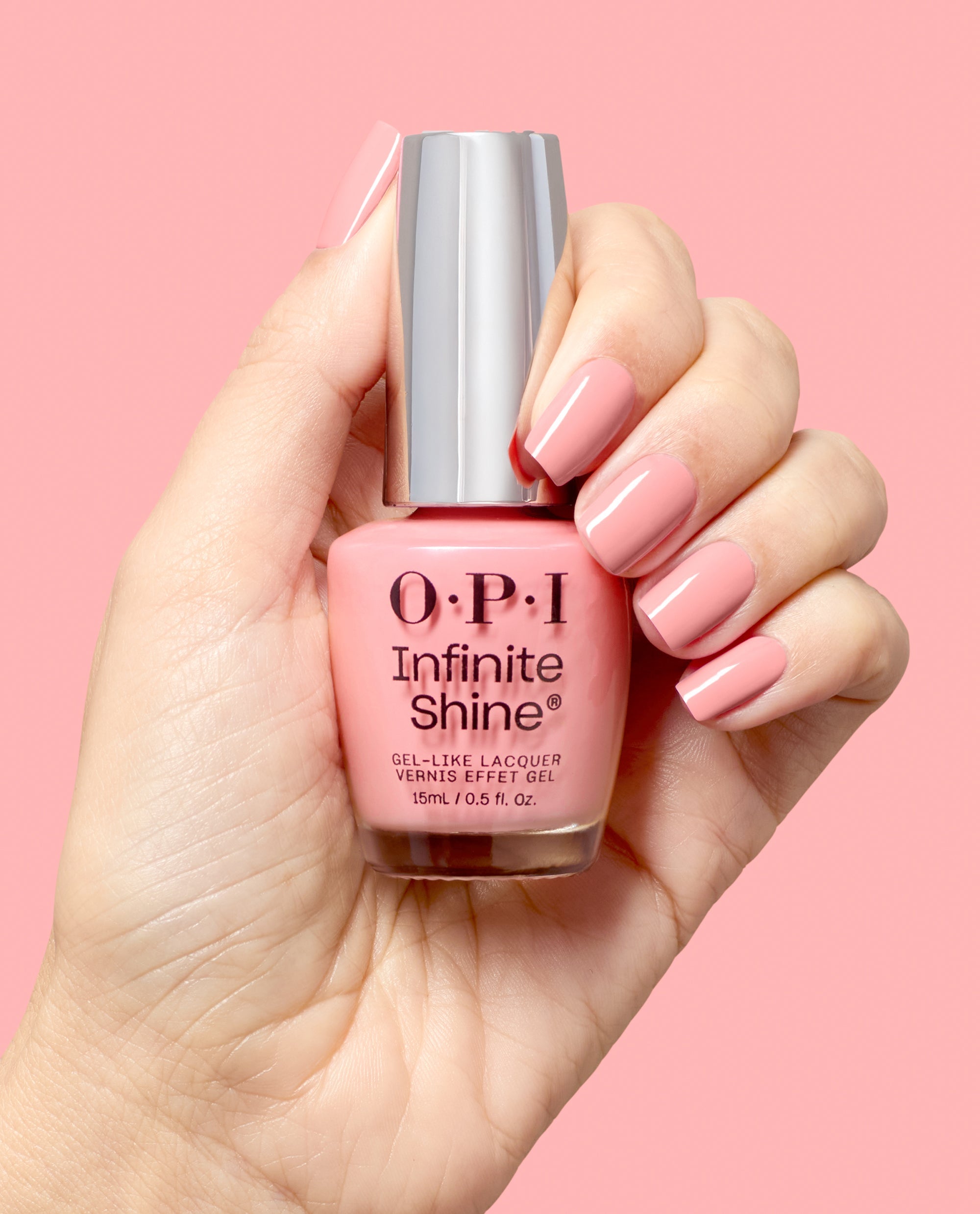 A hand with glossy soft pink manicured nails holds an OPI Infinite Shine Still Going Strong gel-like lacquer bottle against a pink background, showcasing a polished fall nail color.
