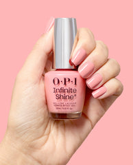 A hand with glossy soft pink manicured nails holds an OPI Infinite Shine Still Going Strong gel-like lacquer bottle against a pink background, showcasing a polished fall nail color.