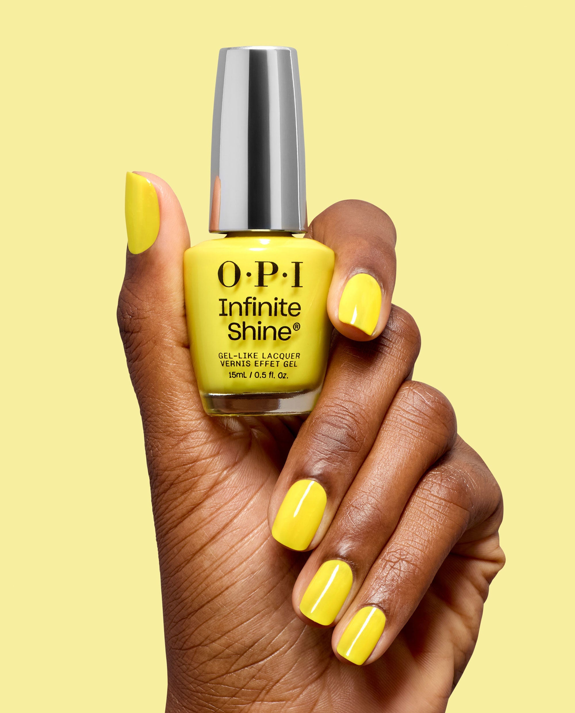 Hand with glossy yellow nails holds OPI Infinite Shine Sunkissed and Tell gel-like lacquer bottle with a silver cap against a pale yellow background.
