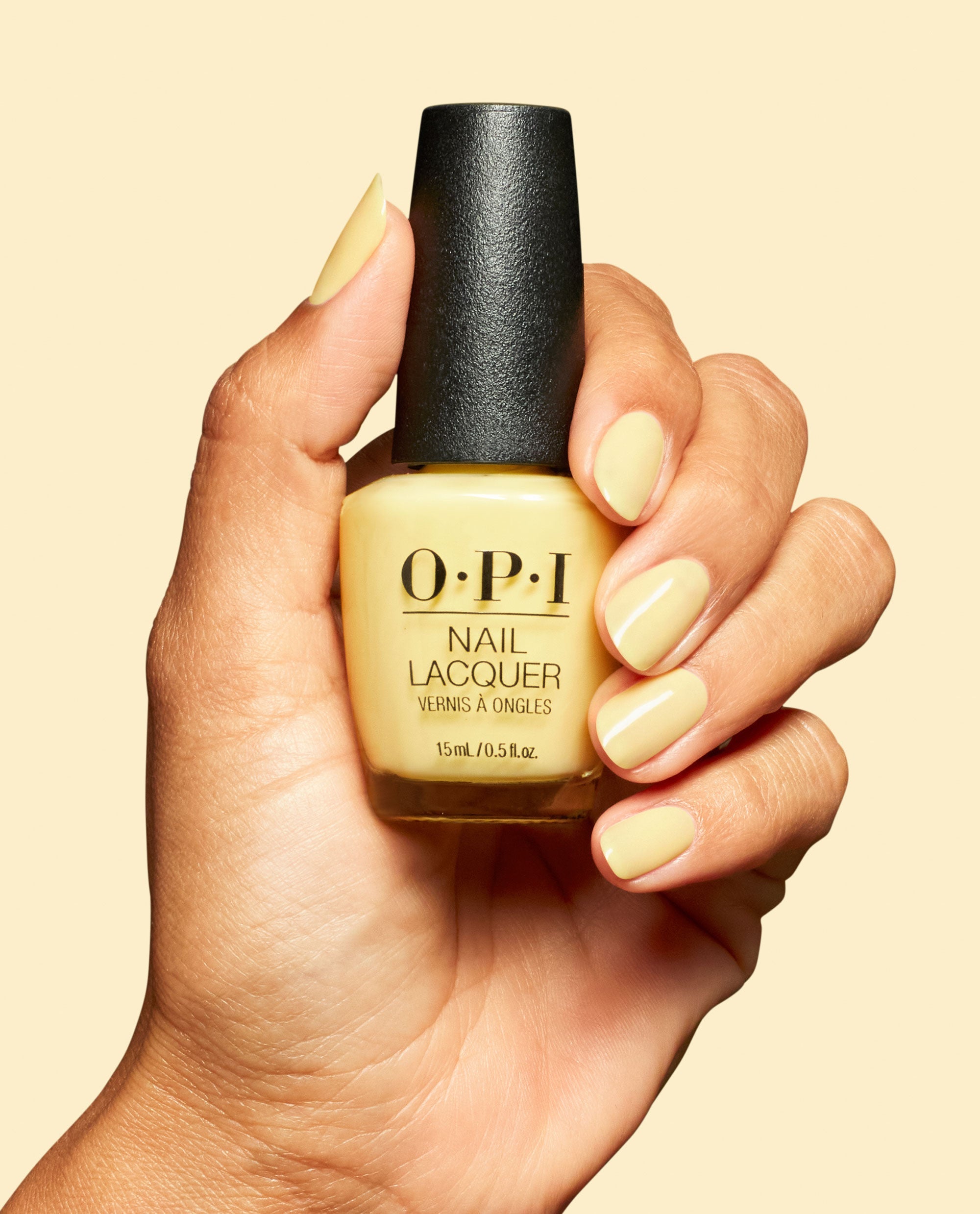 Hand with glossy pale yellow nails holds an OPI Sunny Bunny Nail Lacquer bottle with a black cap against a soft yellow background.