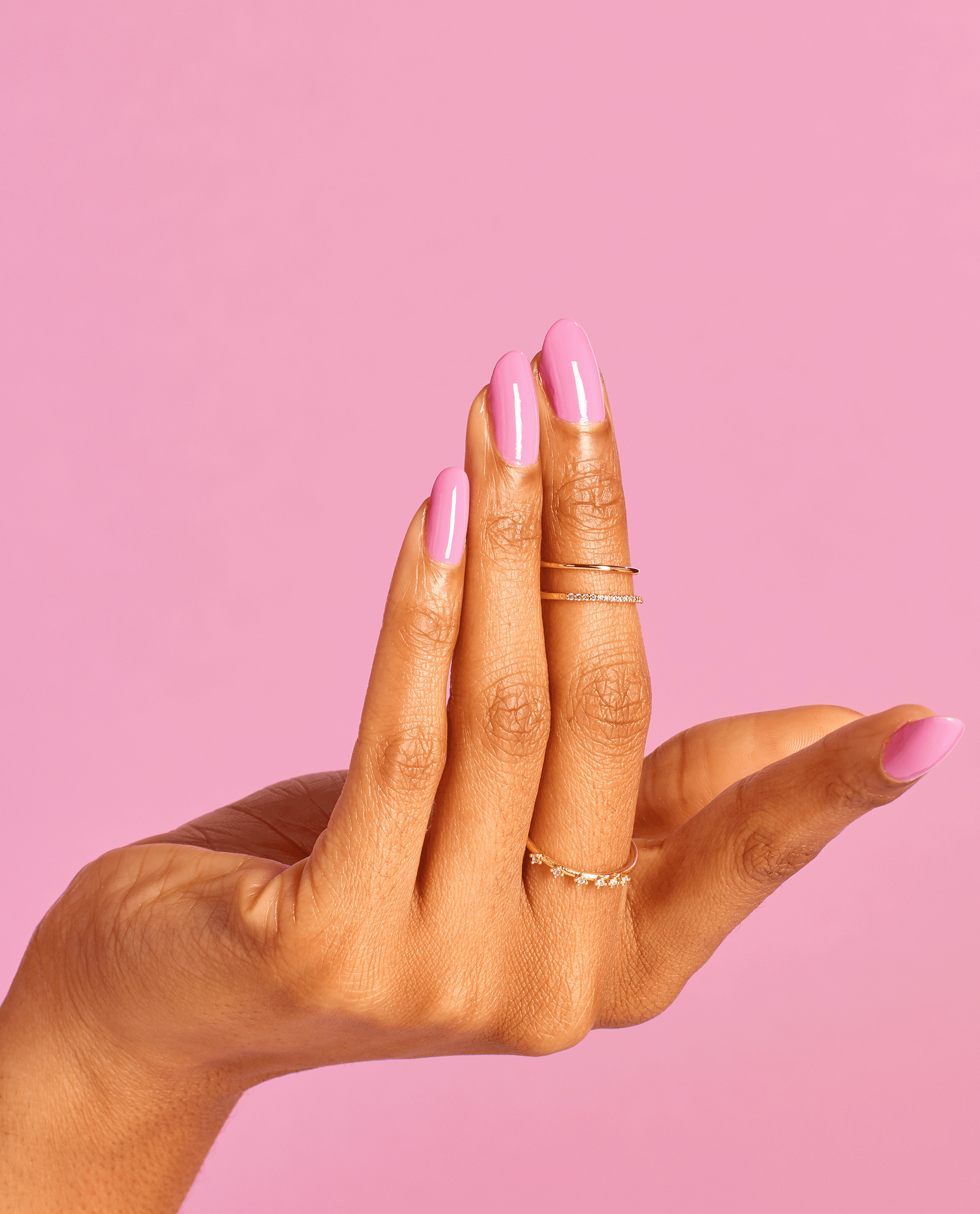 Manicured hand with medium skin tone flexing glossy pastel pink Suzi Will Quechua Later! OPI nail polish, nails oval-shaped and paired with delicate gold rings, against a solid pink studio background showcasing the nail lacquer color and shine.