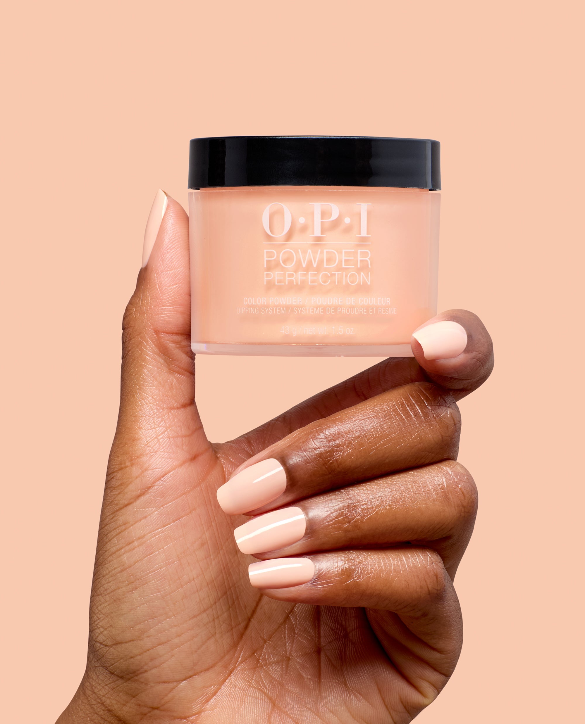 OPI Suzi Take a Vow Powder Perfection dipping powder jar held by a hand with glossy soft peach manicured nails against a peach background, showcasing a neutral shade for fall nail colors.