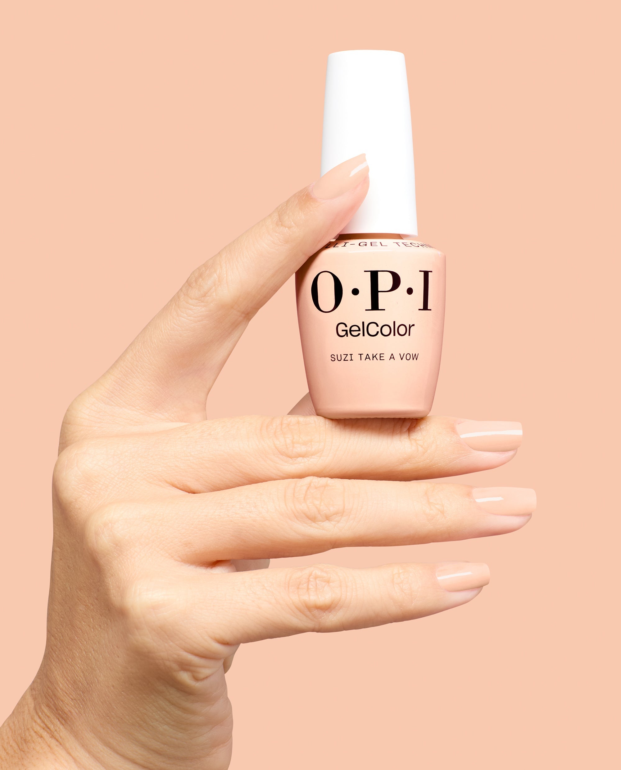 Hand with glossy nude peach manicure holds OPI GelColor Suzi Take a Vow gel nail polish bottle against a matching peach background, a soft neutral pick for fall nail colors.