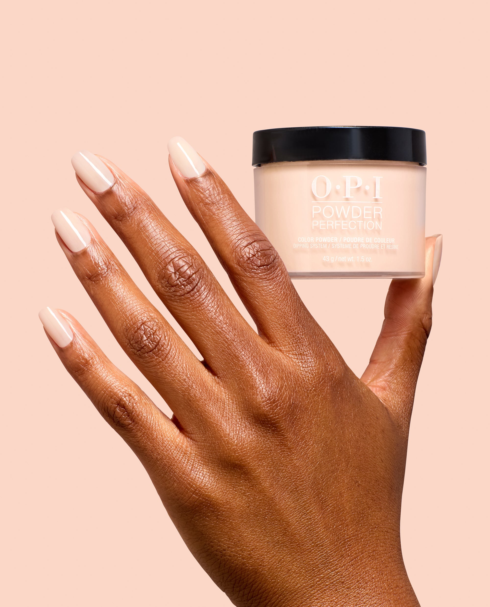 Hand with glossy nude manicure holds OPI Powder Perfection Throw Me a Wink dipping powder jar, a soft neutral shade for fall nail colors, against a peach background.