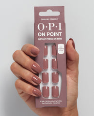 Hand with mauve-pink manicured nails holds OPI On Point Tickle My France-y instant press-on mani package, showing 24 glossy nude press-on nails from an OPI nail polish gift set against a light gray background.