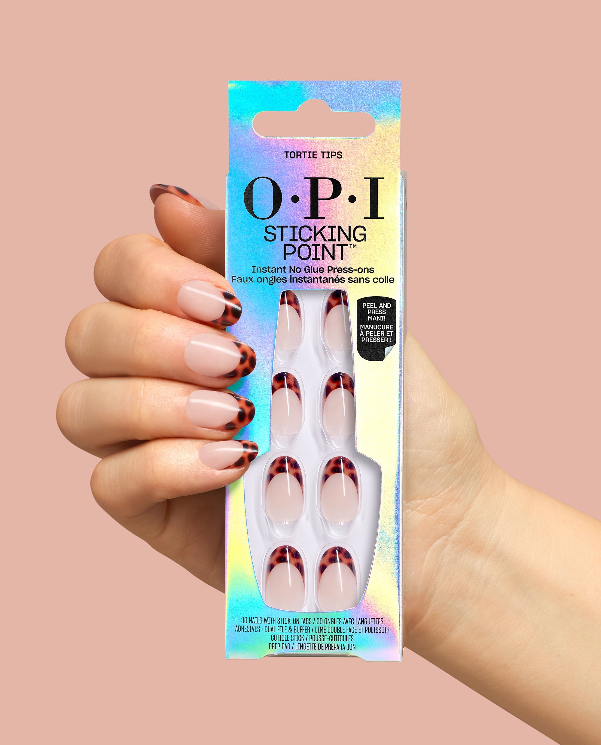 Hand with OPI Tortie Tips press-on nails holds a holographic Sticking Point box, showing nude nails with tortoiseshell French tips for chic fall nail colors on a peach background.