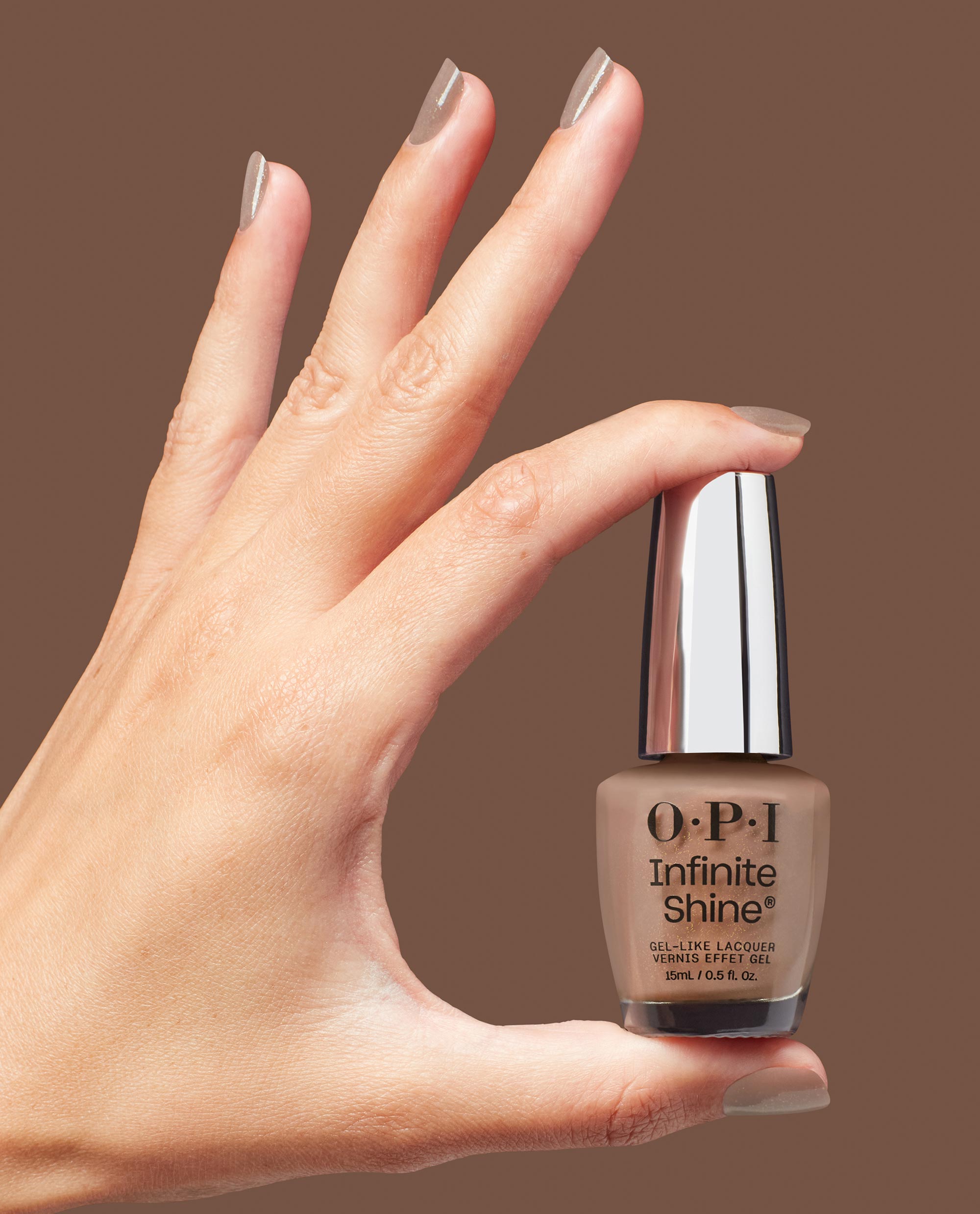 Hand with glossy taupe nails holds OPI Infinite Shine Track Suited Cutie gel-like lacquer bottle, a warm neutral shade for fall nail colors, against a brown studio background.