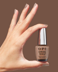 Hand with glossy taupe nails holds OPI Infinite Shine Track Suited Cutie gel-like lacquer bottle, a warm neutral shade for fall nail colors, against a brown studio background.