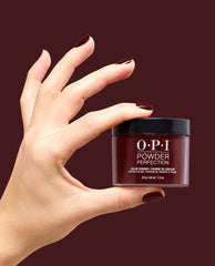Hand with glossy deep burgundy nails holds OPI Powder Perfection Vampire State Building dipping powder, a rich shade for fall nail colors, against a matching wine-red background.