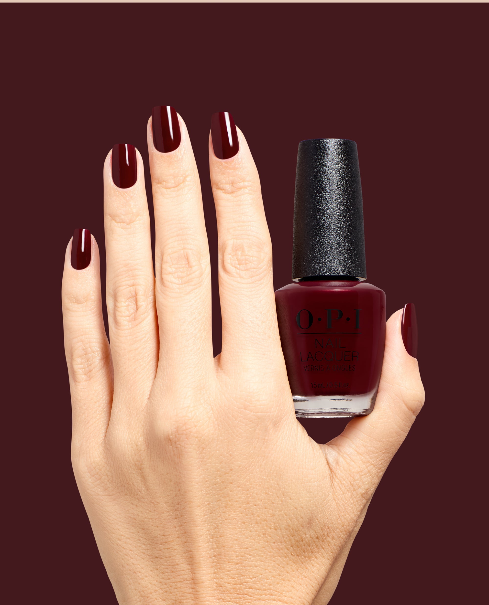 Hand with glossy deep burgundy nails holds OPI Vampire State Building nail lacquer bottle against a matching dark red background, showcasing rich fall nail colors.