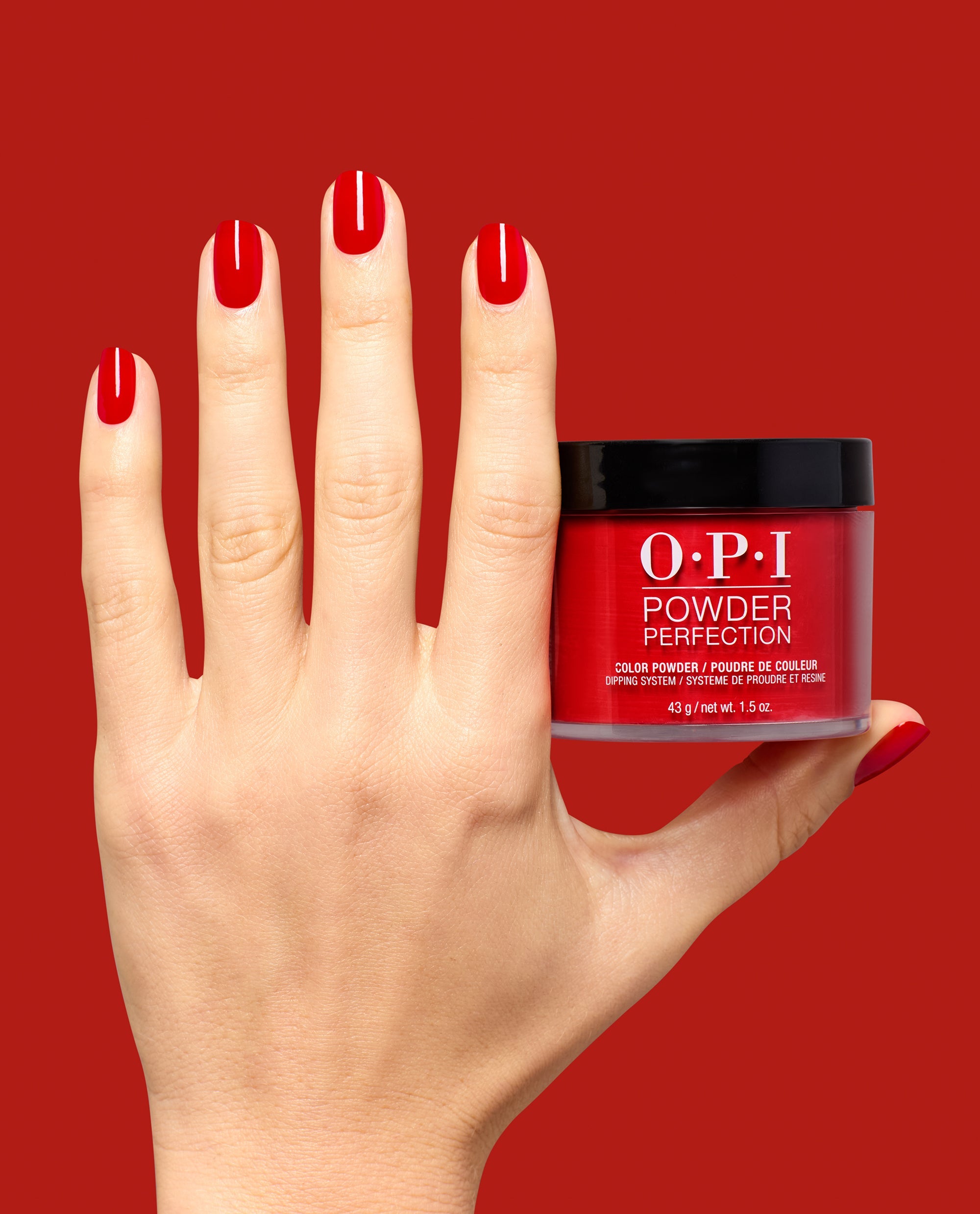 Hand with glossy red nails holds OPI Powder Perfection Vodka & Caviar dipping powder jar against a red background, showcasing a classic shade for fall nail colors.