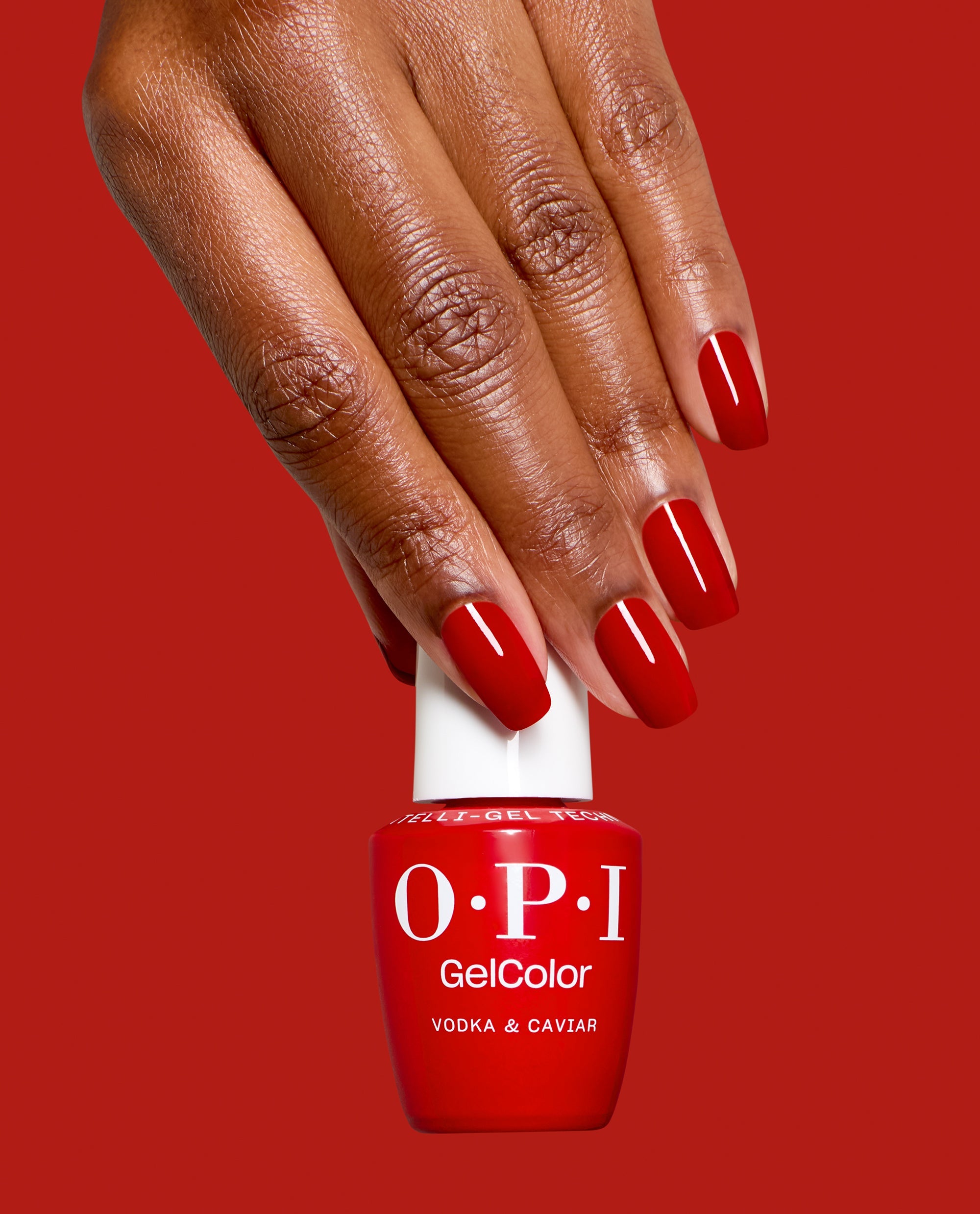 Hand with glossy red nails holds an OPI GelColor Vodka & Caviar gel nail polish bottle against a red background, showcasing a bold classic shade for fall nail colors.