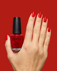 Hand displays glossy red OPI Vodka & Caviar nail lacquer on short square nails while holding the matching bottle against a red background, a bold choice for fall nail colors.