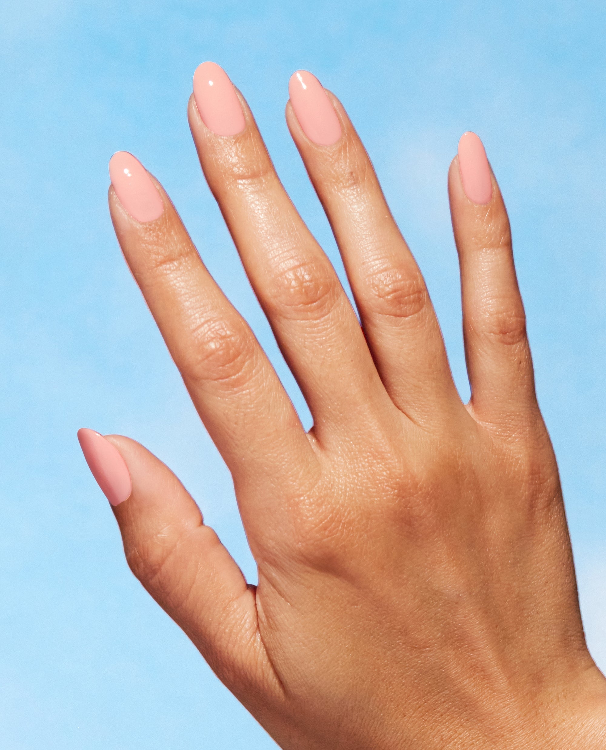Hand with neatly manicured oval nails painted in OPI We Canyon Do Better, a soft peach-pink Nature Strong vegan nail lacquer, shown against a bright blue background for an OPI nail polish gift set product image.