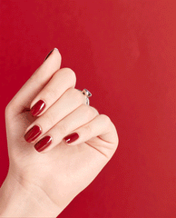 Hand shows glossy deep red OPI We the Female nail lacquer on short rounded nails against a red background, a rich choice among fall nail colors.