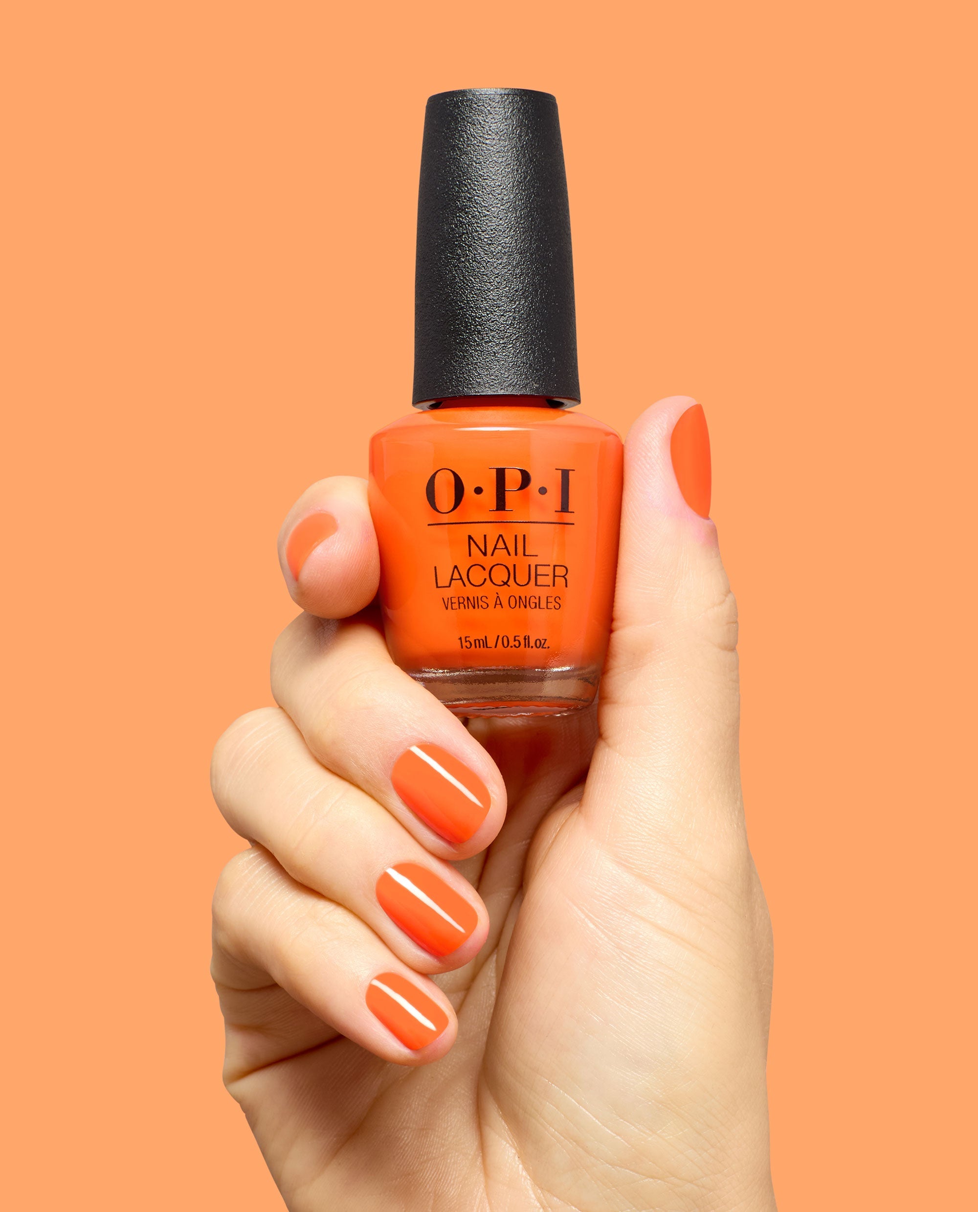 Hand holds OPI Your Zestination Awaits Nail Lacquer in bright orange, with glossy matching orange nails against a peach background.